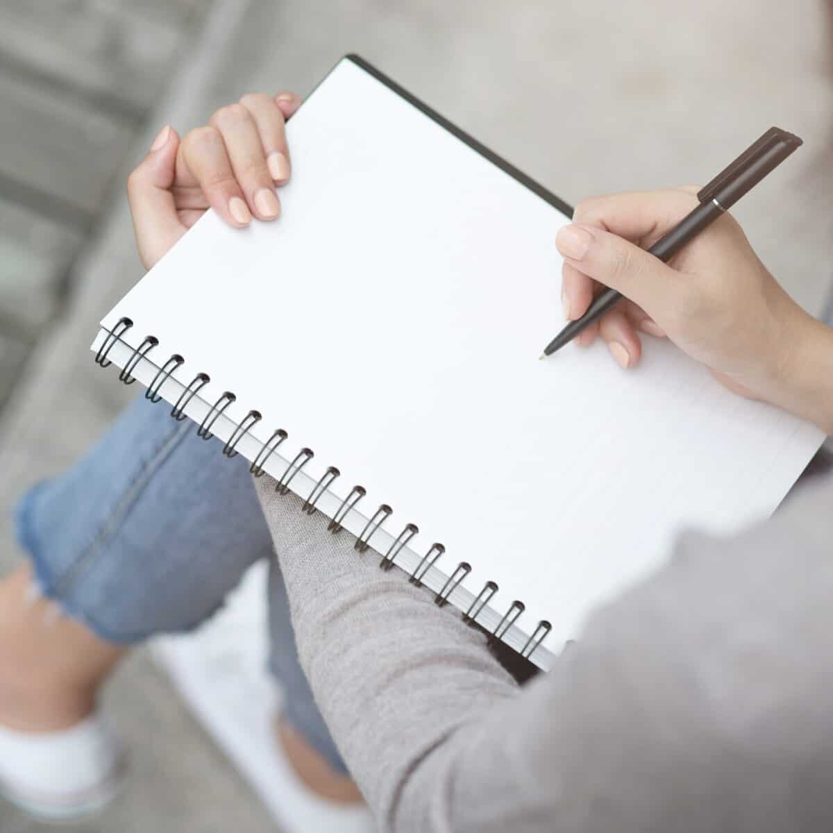 A person sitting with crossed legs and a sketch book on her lap and pencil in hand preparing to practice drawing.